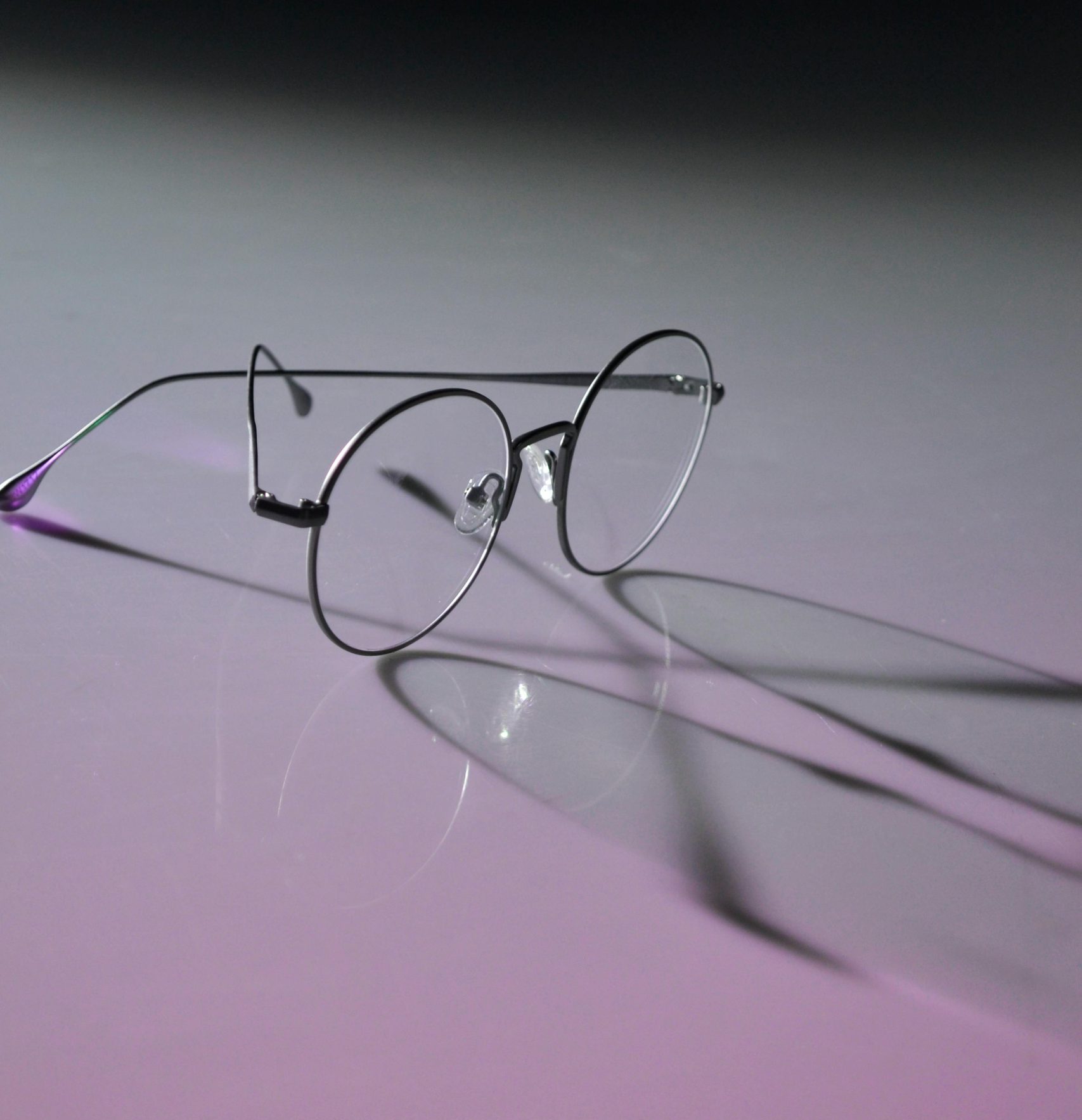 Round eyeglasses with metal frames casting shadows on a neon purple surface, emphasizing modern eyewear style.